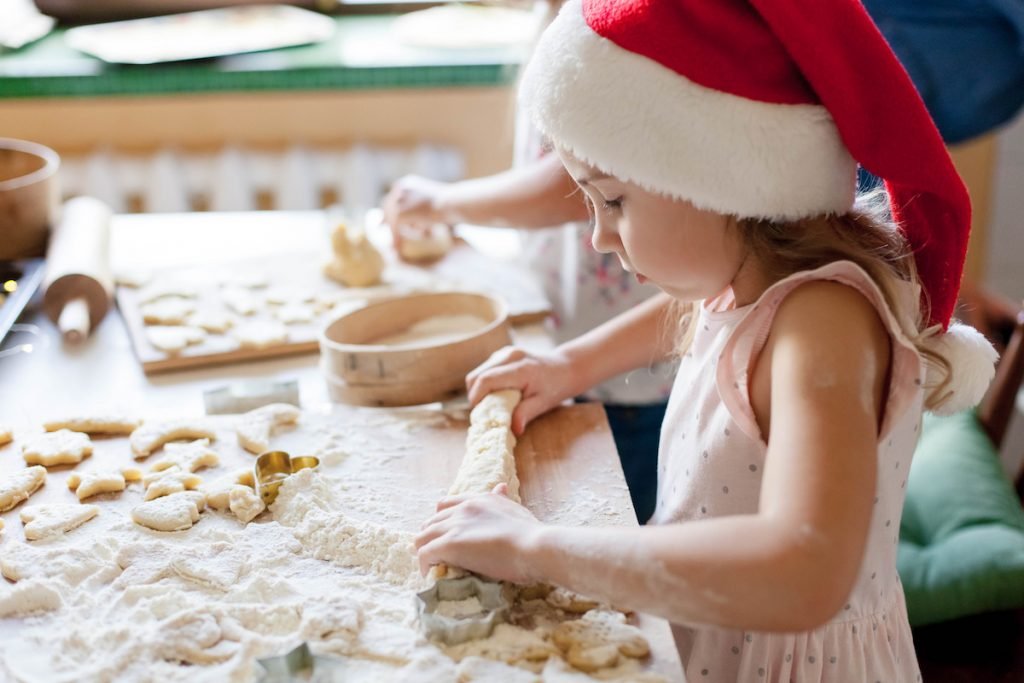 Kids cooking Christmas cookies in cozy kitchen. Child prepares holiday food for family. Cute little girls bake homemade festive gingerbreads. Lifestyle moment. Santa helper. Children chef concept; blog: 10 Holiday Activities The Whole Family Will Love